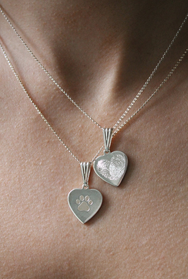 Close-up of two Personalised Charms silver heart pendants on thin chains: one engraved with a paw print, the other with a fingerprint design. They rest elegantly on bare skin.