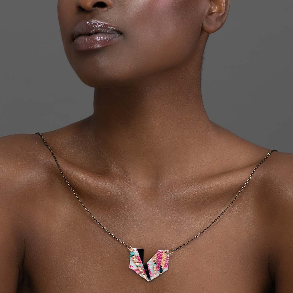 A person with short hair is shown from the shoulders up against a neutral background, wearing the Rothlú Ezrine Batik Textile Necklace in Pink/Rose-Gold/Blue/Black. Their lips are glossed, and their shoulders are bare.