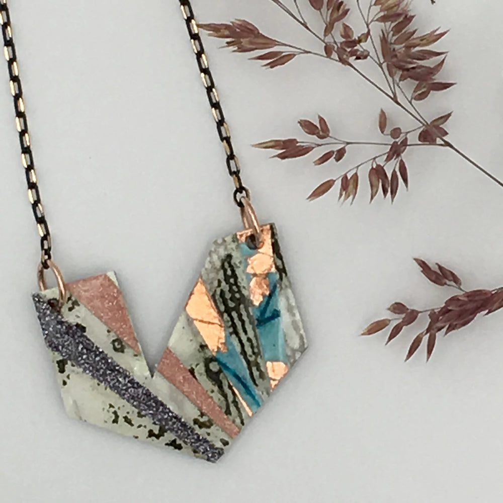 The Ezrine Batik Textile Necklace by Rothlú is a unique piece crafted from two irregularly shaped components, suspended on a black and gold chain. This one-of-a-kind pendant showcases a stunning mix of colors and textures, including olive, white, blue, rose-gold, blush, and pewter shimmer. Decoratively arranged beside the necklace are dried leaves with a reddish hue.