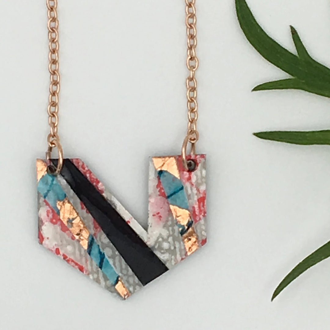 The Ezrine Batik Textile Necklace by Rothlú, featuring pink, rose gold, blue, and black hues, is crafted from a geometric piece of eco resin-coated folded paper. The hand-formed pendant hangs elegantly from a gold chain and is displayed against a white background with green leaves nearby.