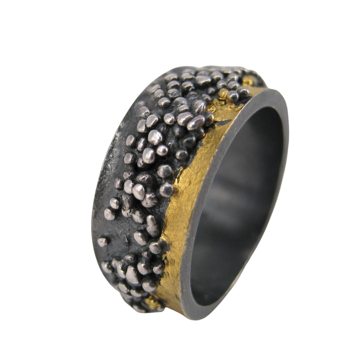 A close-up of a textured ring from the MarCucurella Etrusc Ring collection, featuring a rugged design with a mix of silver and gold elements. The silver part has a rough, clustered pattern reminiscent of Etruscan granulation, while the gold part appears smooth. The wide band and unique style give it an artisanal, handcrafted look.