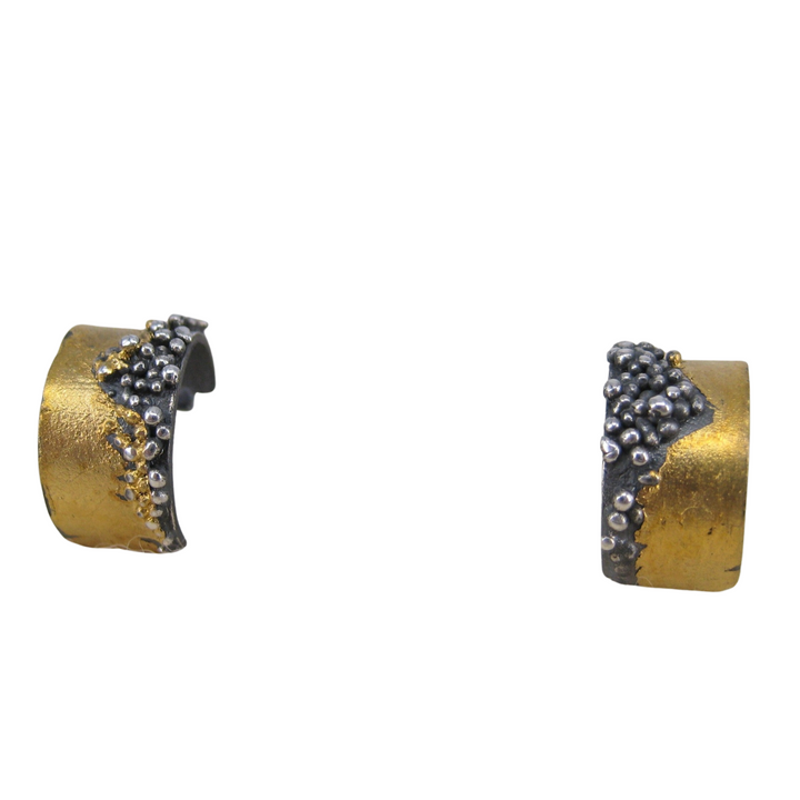 A pair of Etrusc Small Hoop Earrings by MarCucurella. The earrings feature an oxidized silver base with a partially 24 kt yellow gold-toned surface. They are adorned with small clusters of silver and gold beads, creating a textured and artistic design reminiscent of Etruscan granulation techniques.