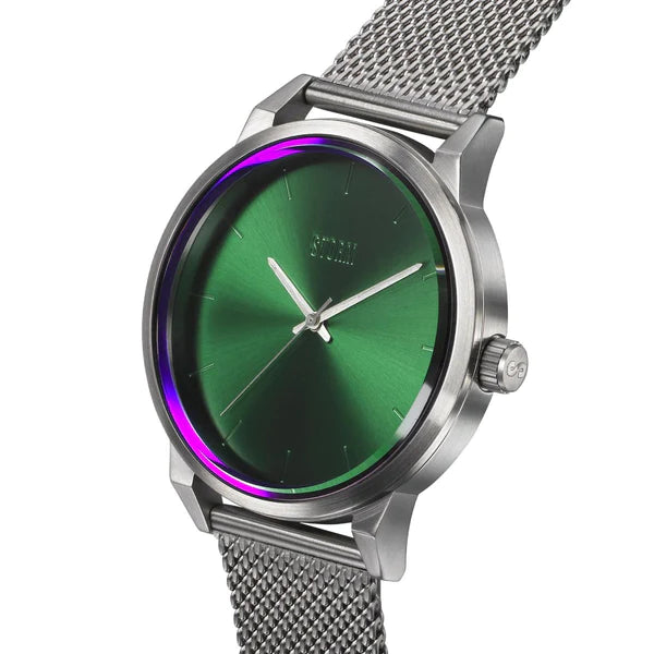 A close-up of a stylish, minimalist design watch with a metallic mesh band. The watch face is green with a subtle radial gradient and features thin silver hour markers and hands. Made from sustainable materials, the modern metallic case complements the purple-hued crystal reflections beautifully. This is the Storm Excepto Green by Storm Watches.