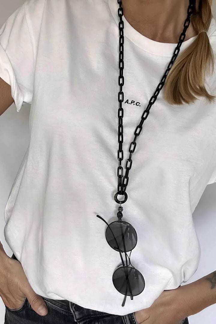 A person wearing a white T-shirt with "A.P.C." printed on it has an AnnaeAlex EyesOnOff Eyewear Black Paperclip Chain around their neck, from which sunglasses are hanging. The person's hand rests on their hip, and they have a side ponytail. The background is plain white.