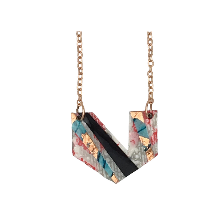 The Ezrine Batik Textile Necklace by Rothlú features a gold chain and a hand-formed pendant with an abstract, multi-colored design in pink, rose-gold, blue, and black. The pendant is uniquely shaped in a folded, angular "V," showcasing varied patterns on each segment. It also boasts a distinct texture and an eco resin finish.