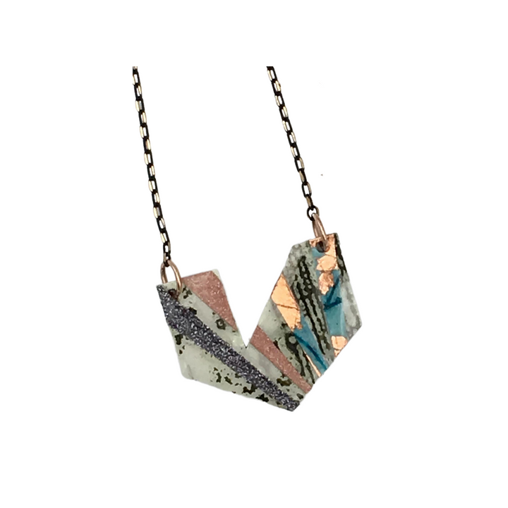 The Ezrine Batik Textile Necklace by Rothlú features a geometric pendant with angular, interlocking shapes and a mix of abstract patterns in olive, white, blue, rose-gold, blush, and pewter shimmer accents. This unique textured necklace is suspended from a delicate metal chain.