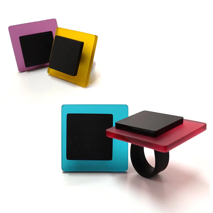 A photo of three Eriko Rings with black bands and colorful, transparent bases. One ring is red with a blue square, another is yellow with a black square, and the last is purple with a black square. The contemporary touch of these Alex + Svet INFINITE LINE collection pieces stands out against the white background.