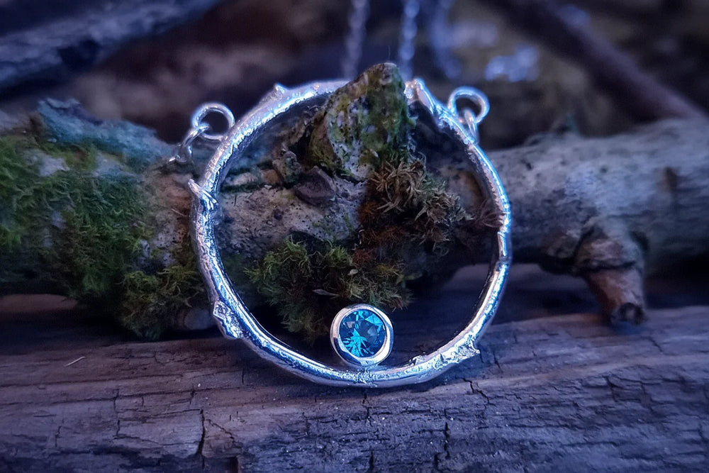 The Faerie Ring Pendant in Blue Topaz by Caroline Stokesberry-lee features a silver circular design with twig-like texture, encasing a blue topaz gemstone at its base, shown atop mossy, weathered wood.