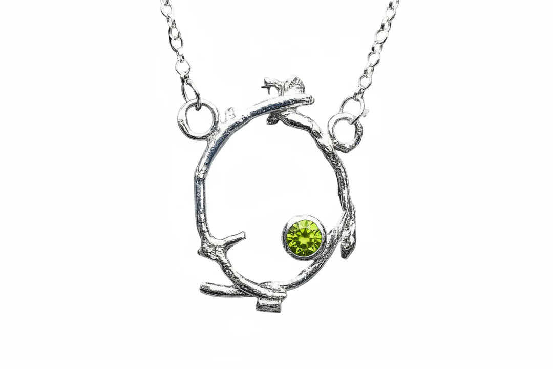 The Faerie Crown Pendant Peridot by Caroline Stokesberry-lee features a circular twig-shaped silver pendant with a round Peridot gemstone, hanging from two loops on a delicate sterling silver chain.