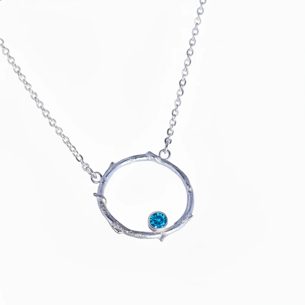 The Faerie Ring Pendant in Blue Topaz by Caroline Stokesberry-lee is a silver necklace with a circular, twig-inspired pendant, featuring a small blue topaz gemstone at the bottom. Displayed on a plain white background.