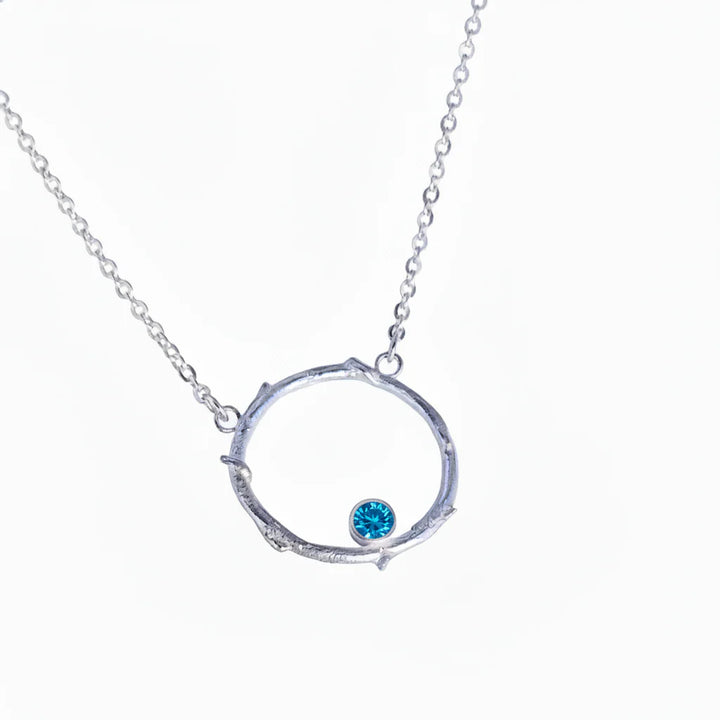 The Faerie Ring Pendant in Blue Topaz by Caroline Stokesberry-lee is a silver necklace with a circular, twig-inspired pendant, featuring a small blue topaz gemstone at the bottom. Displayed on a plain white background.