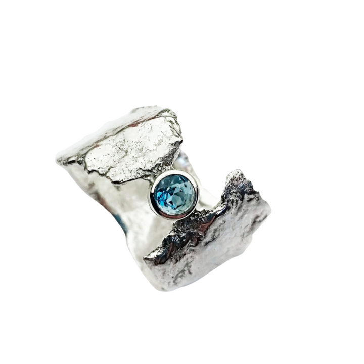 A textured, silver ring with a rugged design features a single, brilliant blue topaz gemstone set prominently on the band. The ring's rough and uneven surface, achieved through reticulation technique, gives it an ethereal faerie tale appearance. The plain white background highlights the ring's details. This is the Faerie Tale Ring In Blue Topaz by Caroline Stokesberry-lee.