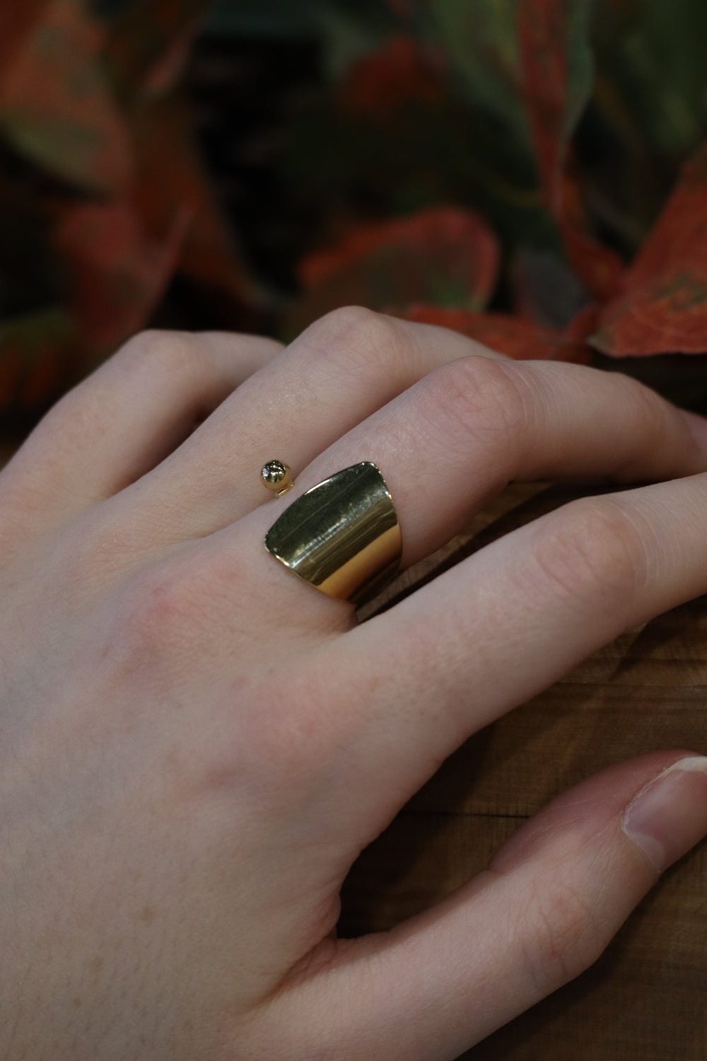A hand rests on a wooden surface, wearing the Zubeldia Gold Plated Curve Ring on the index finger, while blurred reddish-green leaves appear in the background.
