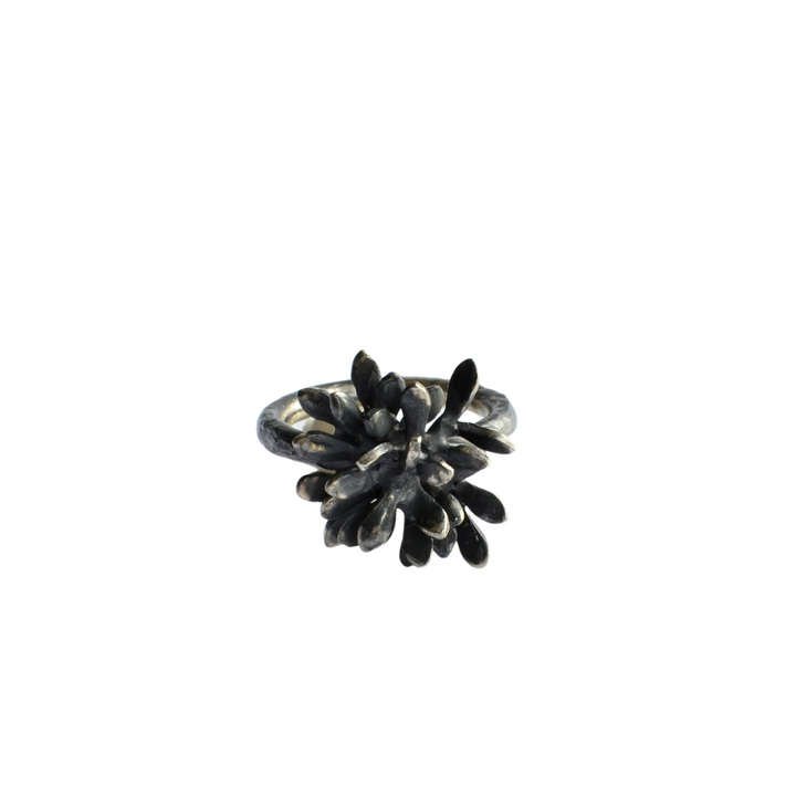 The Folia Oxidised Silver Ring by Abbie Dixon features an intricate, sculptural form with numerous small, petal-like elements radiating from the center, creating a textured, blossoming flower effect. The ring's darkened, oxidized Sterling silver finish adds an antique, artistic touch.