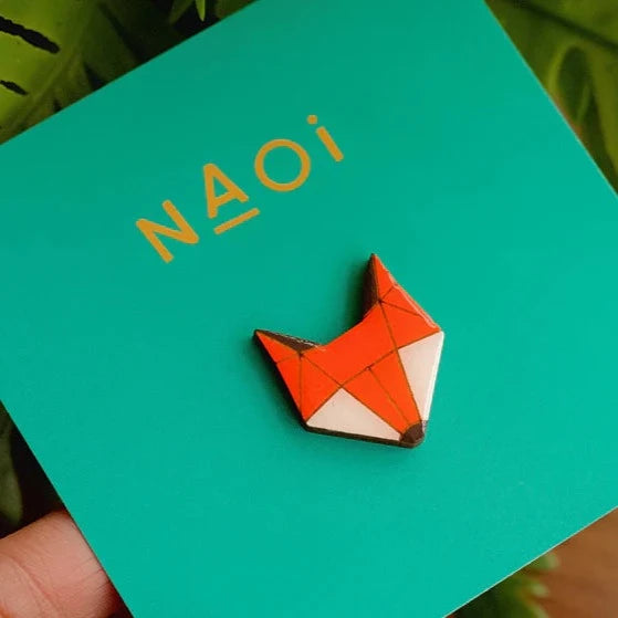 A geometric handpainted wooden Fox Pin Brooch adorns a teal square card with the brand name "Naoi" printed in gold at the top. The Fox Pin Brooch, featuring an orange body, white cheeks, and a black nose, is complemented by a sturdy brass butterfly pin closure.