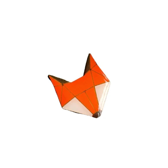 A minimalist, polygonal representation of a fox head. The design, reminiscent of a handpainted wooden fox, features orange, white, and black geometric shapes that form the sharp, angular features of the Fox Pin Brooch by Naoi.