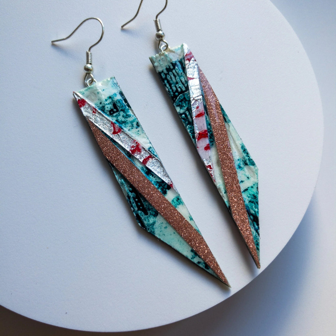 A pair of long, geometric earrings with an abstract design hang on a white background. These unique handmade jewelry pieces feature a mix of teal, white, red, and gray colors with a diagonal glittery bronze stripe. They have sterling silver earring wires for wearing. The product shown is the Foxtrot Batik Textile Earrings in Jade/Coral/Silver and Blush Shimmer by Rothlú.