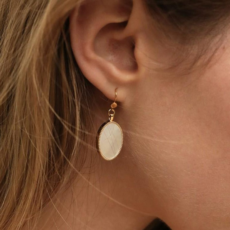 Close-up of a person wearing Nilai's Frida Earrings in an oval shape, featuring a natural gemstone encased in a detailed 24K gold-finished border. Handmade in Paris, this piece beautifully complements the person's ear and light brown hair, which stand out against the softly blurred background.