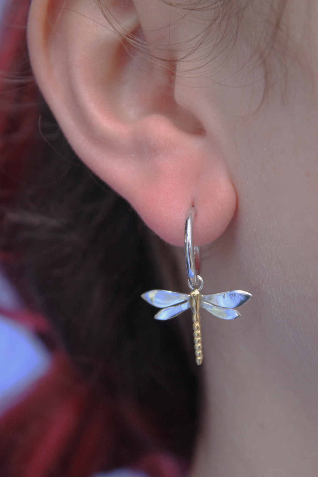 A close-up shows a person wearing Gallardo & Blaine Designs’ Wildlife Dragonfly Dangle Earrings (Silver or Gold), with part of their red hair visible and a blurred background, highlighting this symbolic jewellery piece.