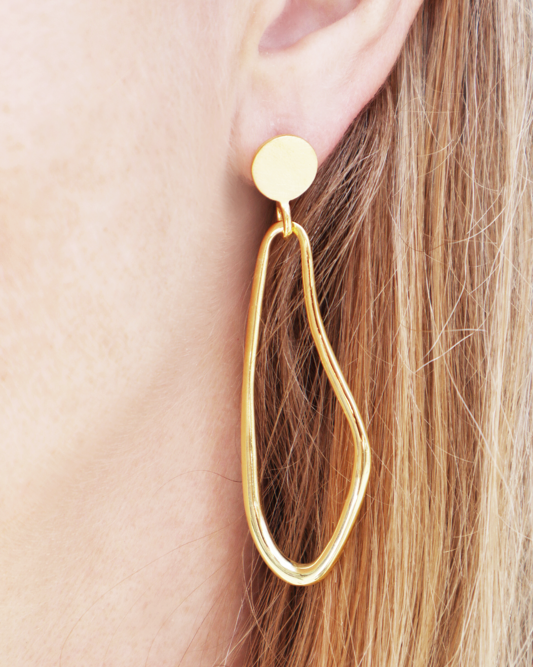 Close-up of a woman's ear wearing Gallardo & Blaine Designs' Sea Honey Drop Earrings, showcasing large abstract gold vermeil hoops with round studs and an organic, irregular shape. Blonde hair is partly visible.