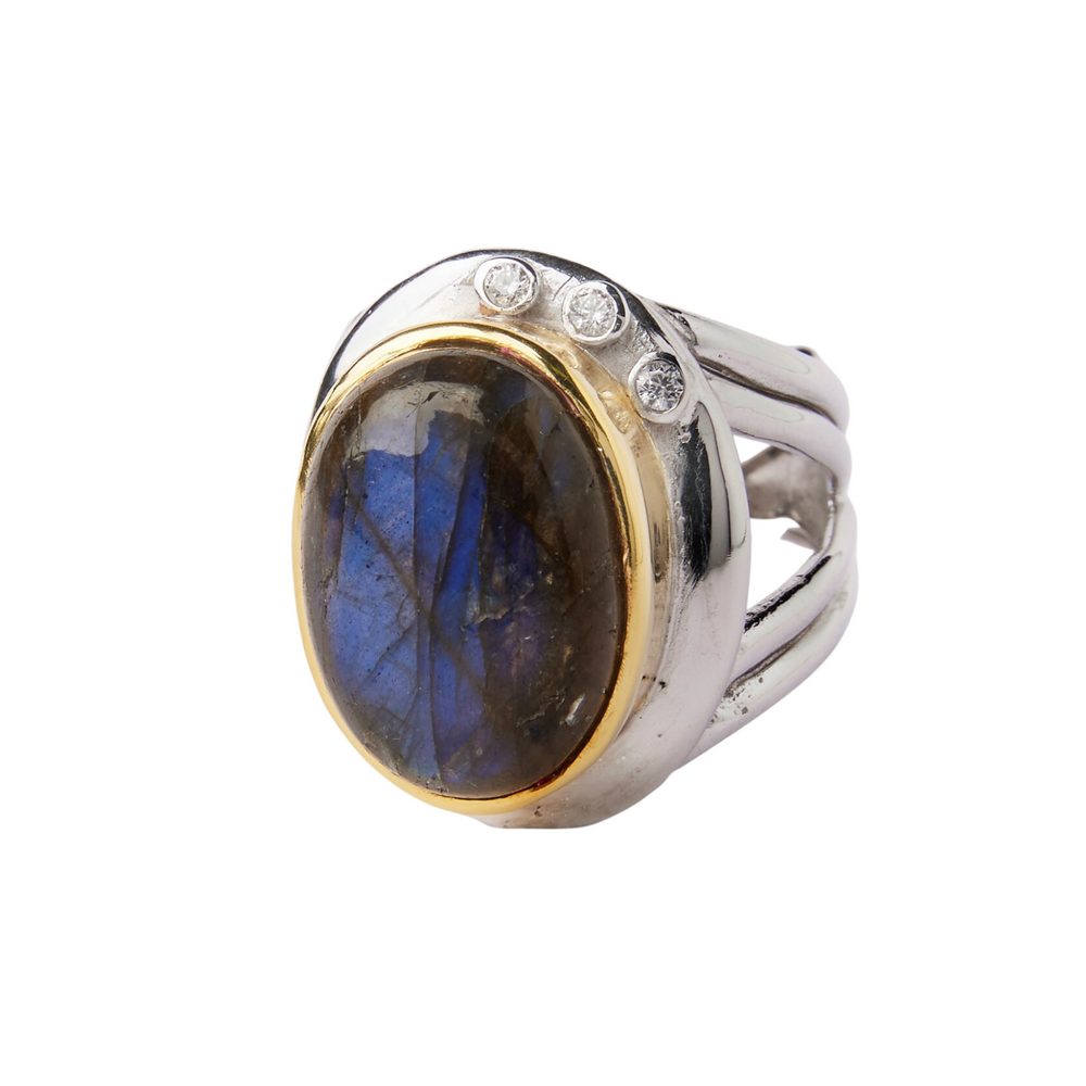 The Eyetelia Ring in various gemstones by Gallardo & Blaine Designs features an oval-shaped labradorite stone, encased in a golden bezel setting with three small diamonds on the top side of the stone. This statement cocktail ring boasts a bold, chunky design with a split shank band.