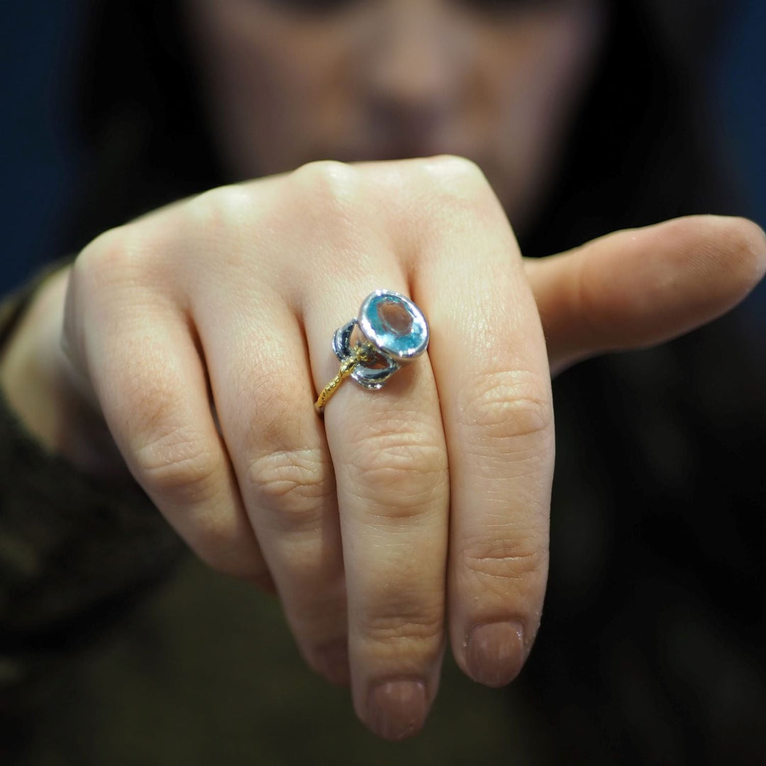 A person wearing a delicate and dainty Gallardo & Blaine Designs Daydream Ring in various gemstones with a large blue gemstone on their finger. The hand is shown up close, with the fingers partially curled and extending towards the camera, making the timeless piece prominently visible. The background is blurred and dark.