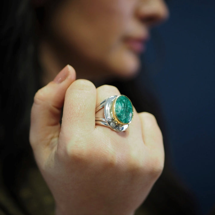 A person displays a Gallardo & Blaine Designs Eyetelia Ring in various gemstones with a large, polished green gemstone. The hand and ring are in focus, while the person's face is blurred in the background. The background is a solid, dark color.