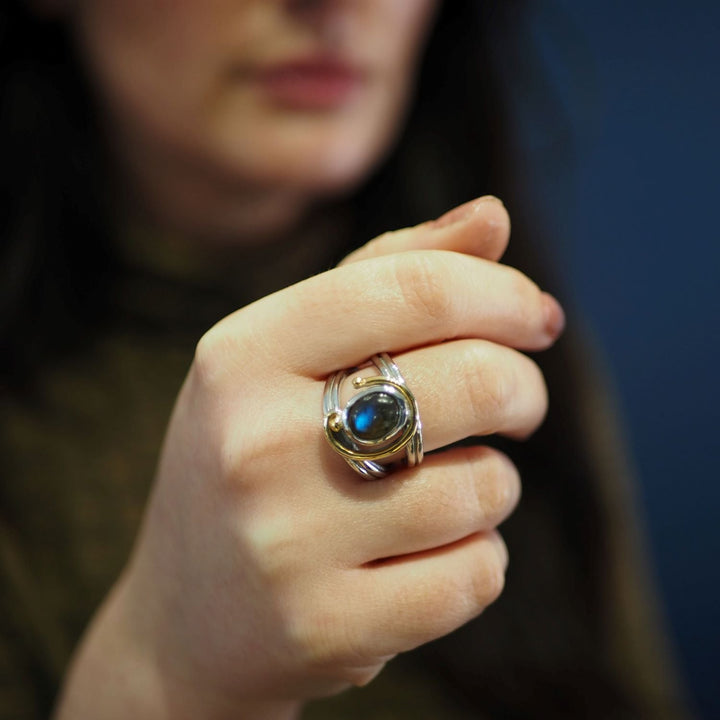 A person with long, dark hair is showcasing a stunning Gallardo & Blaine Designs Jasmine Ring in various gemstones with an adjustable design, featuring a dark, oval gemstone, possibly a blue or black stone. The background is blurred, highlighting the exquisite ring.