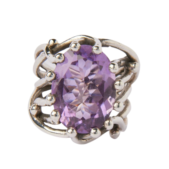 The Willow Ring in various gemstones is a chunky statement gemstone ring crafted in silver with an intricate, wavy design. It features a large, faceted purple gemstone set at the center. Small silver balls accent the design around the adjustable ring, giving it an elegant and ornate appearance. This stunning piece is designed by Gallardo & Blaine Designs.