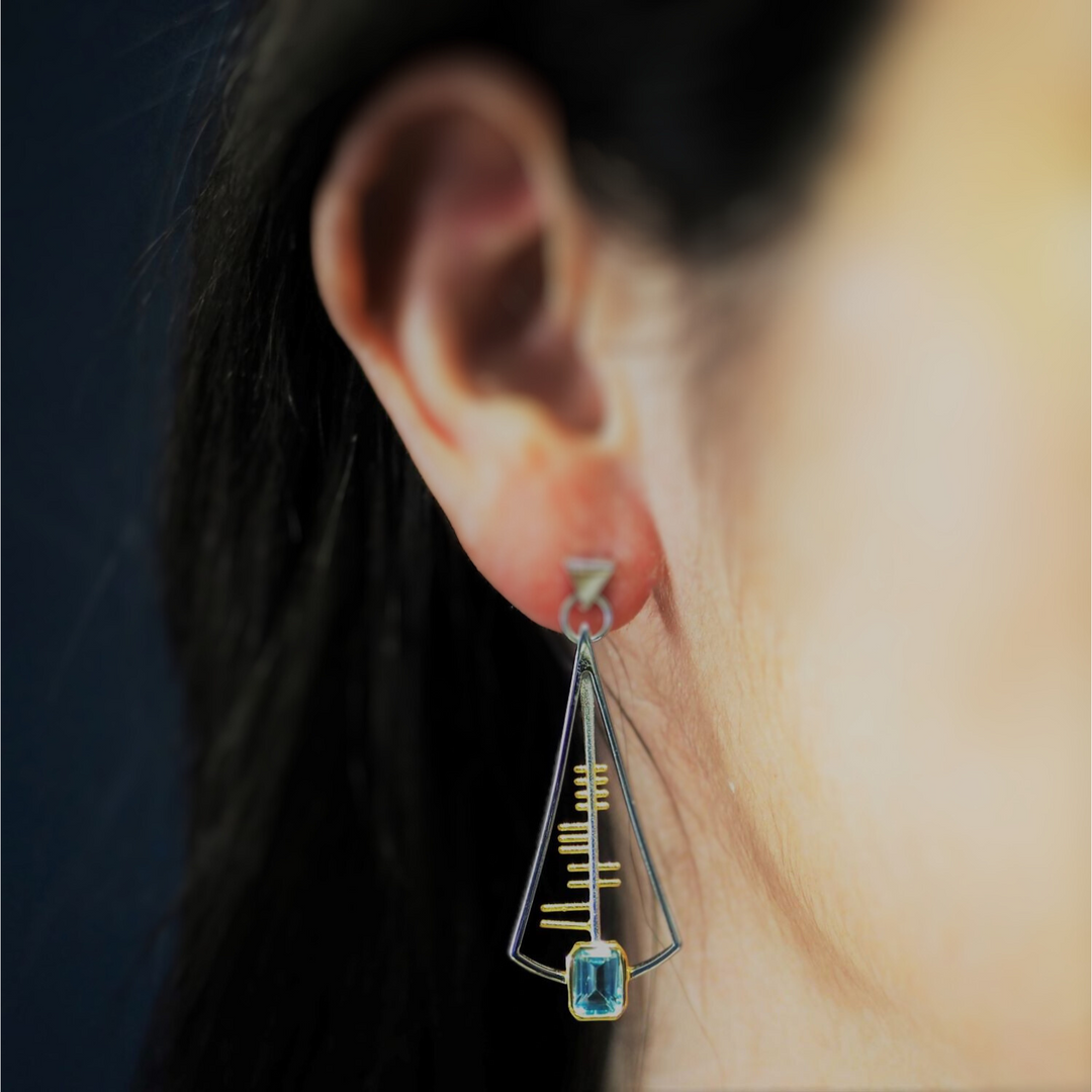 Close-up of a person's ear wearing one of Gallardo & Blaine Designs' Adeline Earrings. The sterling silver triangular frame showcases intricate golden horizontal lines and is adorned with a light blue rectangular gemstone at the bottom. The background is out of focus, highlighting the earring's exquisite details.