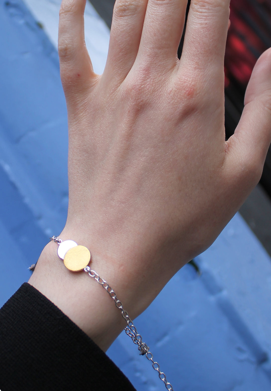 A close-up of a hand wearing Gallardo & Blaine Designs’ Archeol Bracelet in sterling silver and gold vermeil, featuring two round charms inspired by Irish circle jewelry, against a blurred blue background.