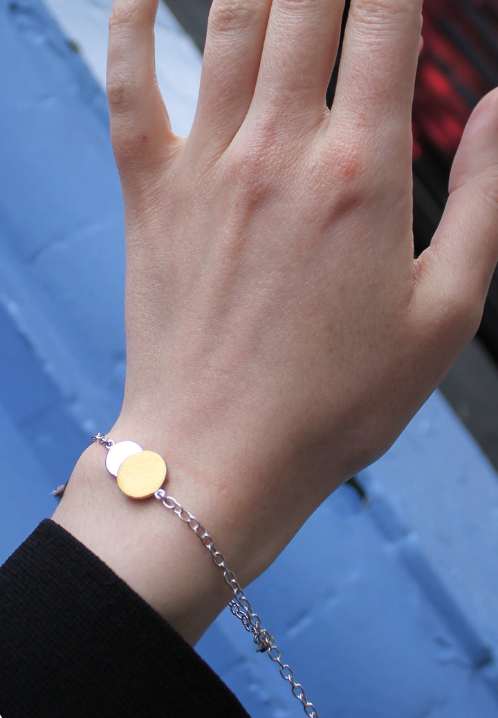 A close-up of a hand wearing Gallardo & Blaine Designs’ Archeol Bracelet in sterling silver and gold vermeil, featuring two round charms inspired by Irish circle jewelry, against a blurred blue background.