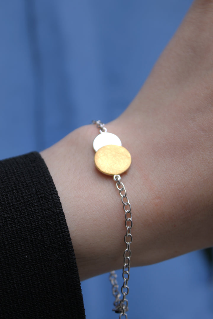 A close-up shows a wrist wearing the Gallardo & Blaine Designs Archeol Bracelet in sterling silver & gold vermeil, featuring two round charms—one gold, one silver—set against blue fabric and a black sleeve.