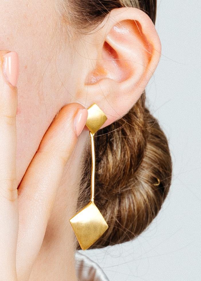 Close-up of a person with a hand touching their face, showcasing an Art Deco earring with a gold vermeil, geometric design from Gallardo & Blaine Designs. The person has light skin and their hair is styled in a low bun. The plain background draws attention to the exquisite Art Deco Earrings In Gold Vermeil by Gallardo & Blaine Designs.