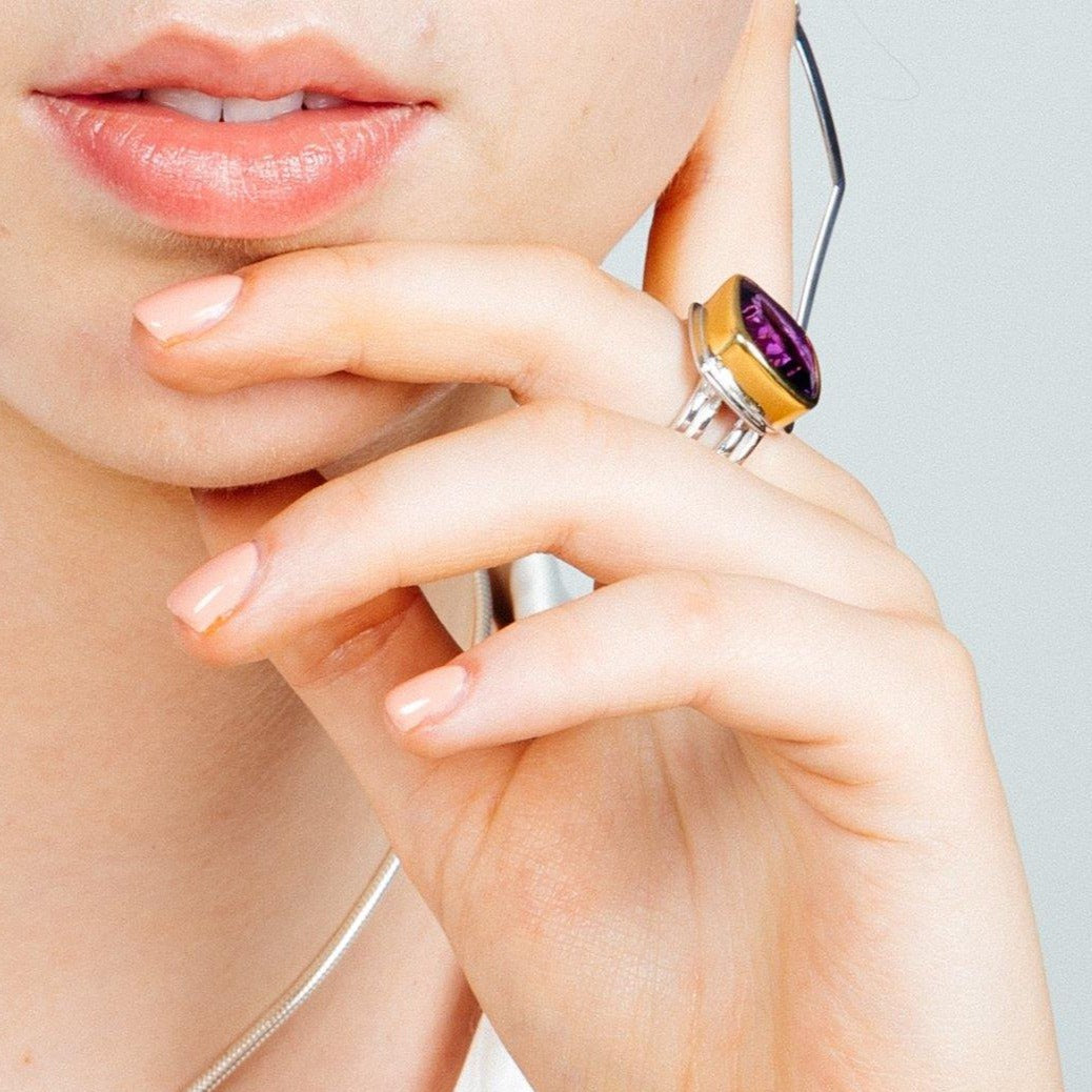 Close-up of a person's face showing their lips and chin, with their hand resting gently on their face. The person is wearing an Art Deco Ring in Silver Gold & various gemstones by Gallardo & Blaine Designs on the index finger and has neatly manicured nails painted a soft peach color.