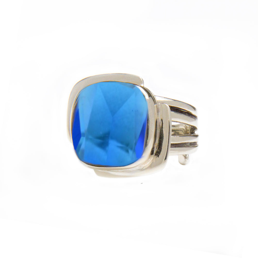 A sterling silver ring featuring a large, faceted blue gemstone. The band has a sleek, modern design with multiple loops for a contemporary look. The square-cut gemstone adds a bold and elegant touch to this Art Deco Ring in Silver Gold & various gemstones by Gallardo & Blaine Designs. The background is plain white.