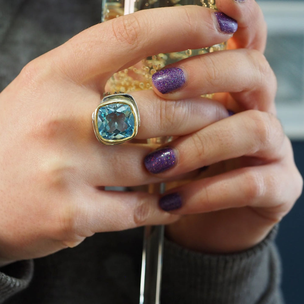 A close-up of a person's hands holding a glass filled with a yellow, bubbly liquid. One hand features glittery purple nail polish and an Art Deco Ring in Silver Gold & various gemstones by Gallardo & Blaine Designs. The background is slightly blurred.