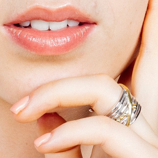 Close-up of a person’s lower face and hand. Their lips are slightly parted, showing teeth. They wear the Bamboo Ring in Sterling Silver & Gold Vermeil by Gallardo & Blaine Designs on their middle finger, featuring intertwined sterling silver and gold vermeil bands. Their fingernails are neatly manicured with a natural, pale pink polish.