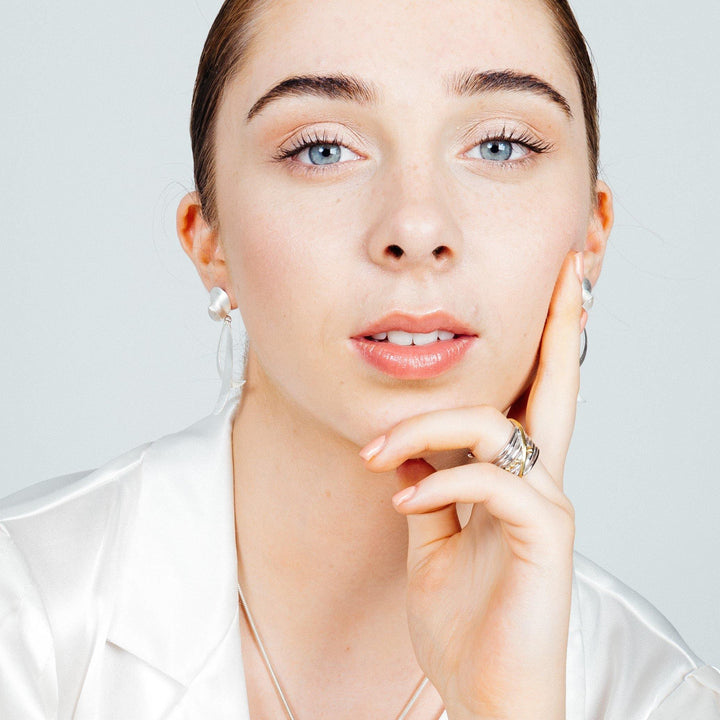 A young woman with fair skin, blue eyes, and brown hair pulled back is gazing into the camera. She is wearing a white top, gold vermeil earrings, and a Gallardo & Blaine Designs Bamboo Ring in Sterling Silver & Gold Vermeil on her index finger. Her hand rests gently on her chin with her fingers touching her cheek.