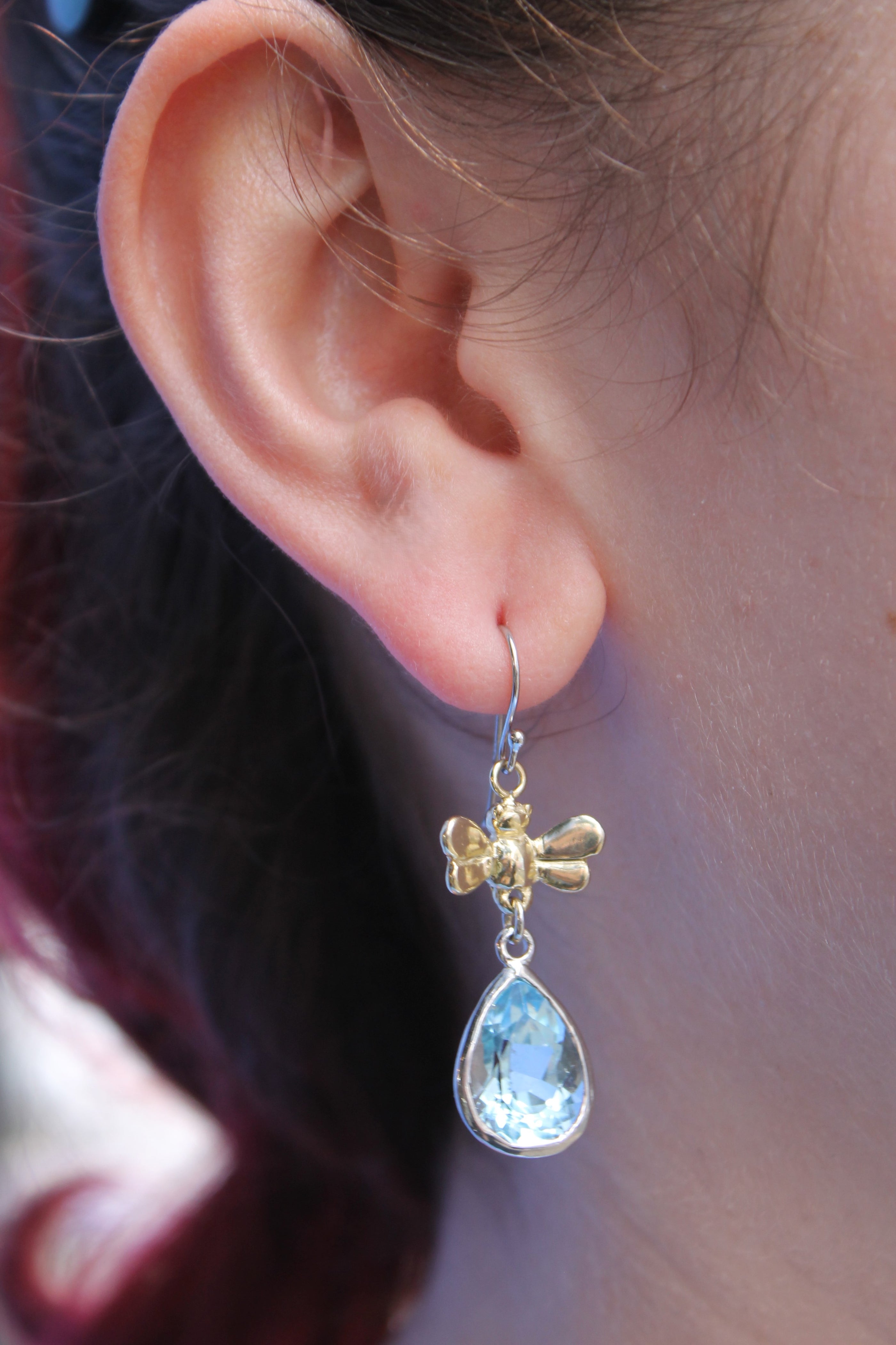 A close-up of a person's ear wearing Gallardo & Blaine Designs' Wildlife Bee Drops, featuring a gold bee and teardrop blue gemstone—nature-inspired earrings for gemstone jewelry lovers.