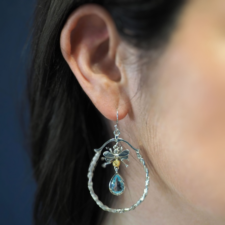 A close-up of a person wearing a sterling silver hoop earring. The intricate design resembles a bee with a blue teardrop-shaped gemstone drop hanging from it. The Wildlife Large Bee Hoop Drops in various gemstones by Gallardo & Blaine Designs dangle elegantly from the person's ear, with their dark hair partially visible.