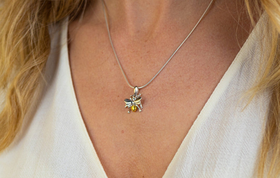 Close-up of a person wearing a delicate sterling silver necklace with a bee pendant, featuring detailed wings and a yellow gemstone. The person is dressed in a light-colored blouse. They are adorning the Wildlife Pendant - Small Bee With Chain by Gallardo & Blaine Designs.