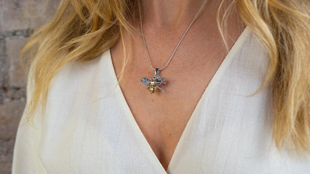 A person wearing a white V-neck shirt with a Gallardo & Blaine Designs Wildlife Pendant - Large Bee With Chain. The person's blonde hair is partially visible against the light, blurred wall in the background.