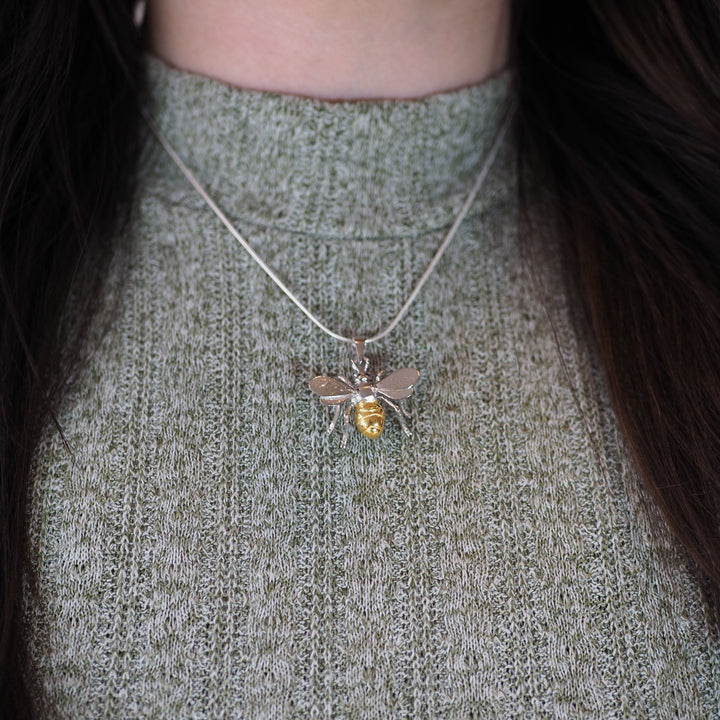 Close-up of a person wearing a grey knit sweater and a silver necklace with the Wildlife Pendant - Large Bee With Chain by Gallardo & Blaine Designs. The Wildlife Pendant - Large Bee With Chain features intricate details, with a gold-plated body and sterling silver wings. The person's long, dark hair frames the necklace, highlighting its delicate design.