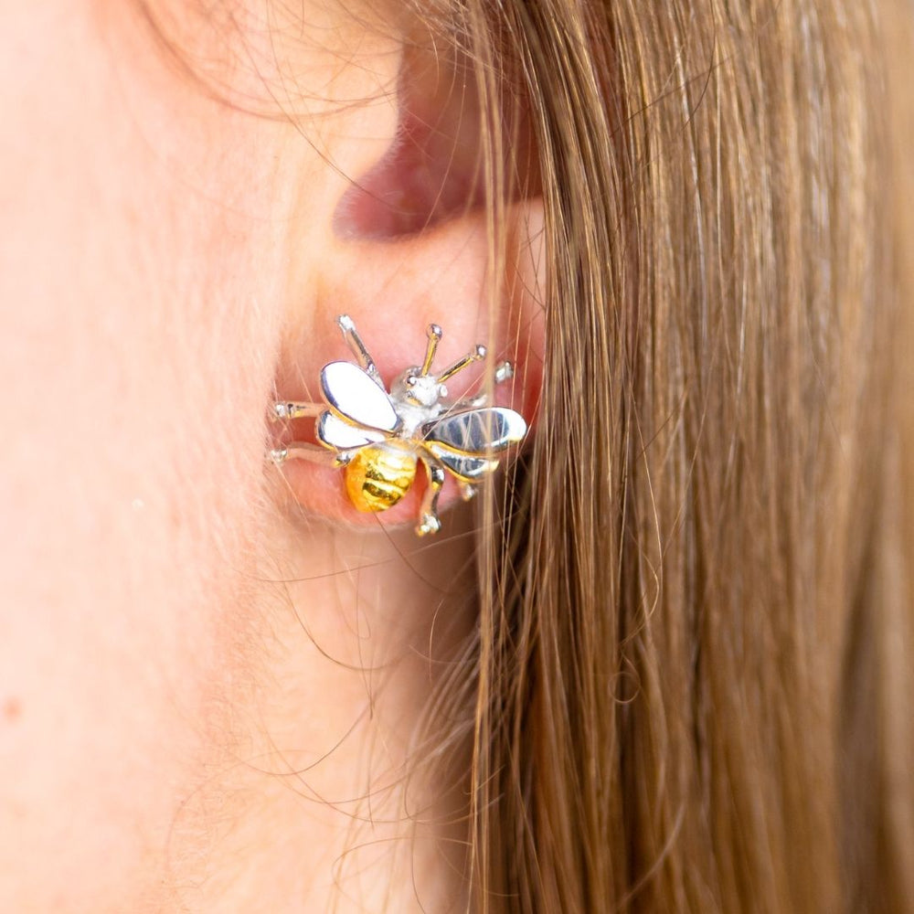 Close-up of a person's ear adorned with Gallardo & Blaine Designs' sterling silver Wildlife Bee Studs from the Wildlife Collection. The earring, with gold plating, is designed to resemble a bee, featuring shiny wings and a detailed body with black and yellow stripes. The person's brown hair is partially visible.