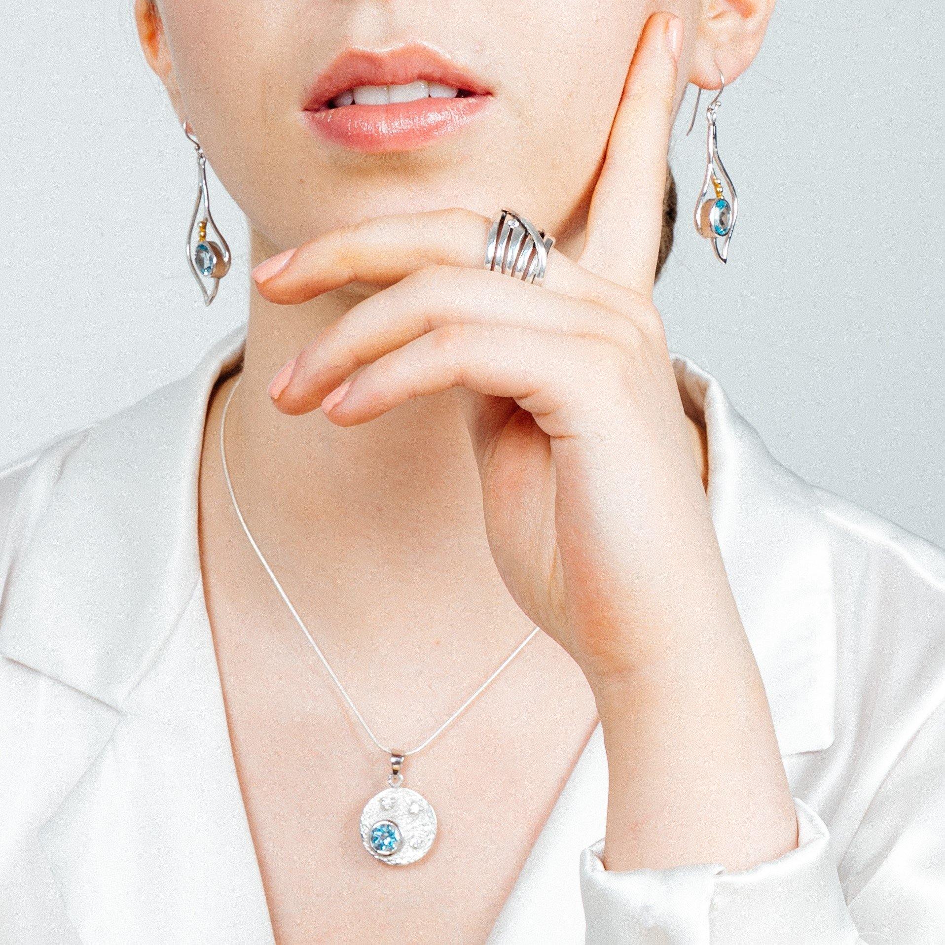 A person wearing elegant jewelry, including dangling earrings with blue stones, a **Gallardo & Blaine Designs Celeste Necklace in Gold & various gemstones**, and a stylish multi-band ring. The person is dressed in a white satin blouse and holds their hand near their chin.