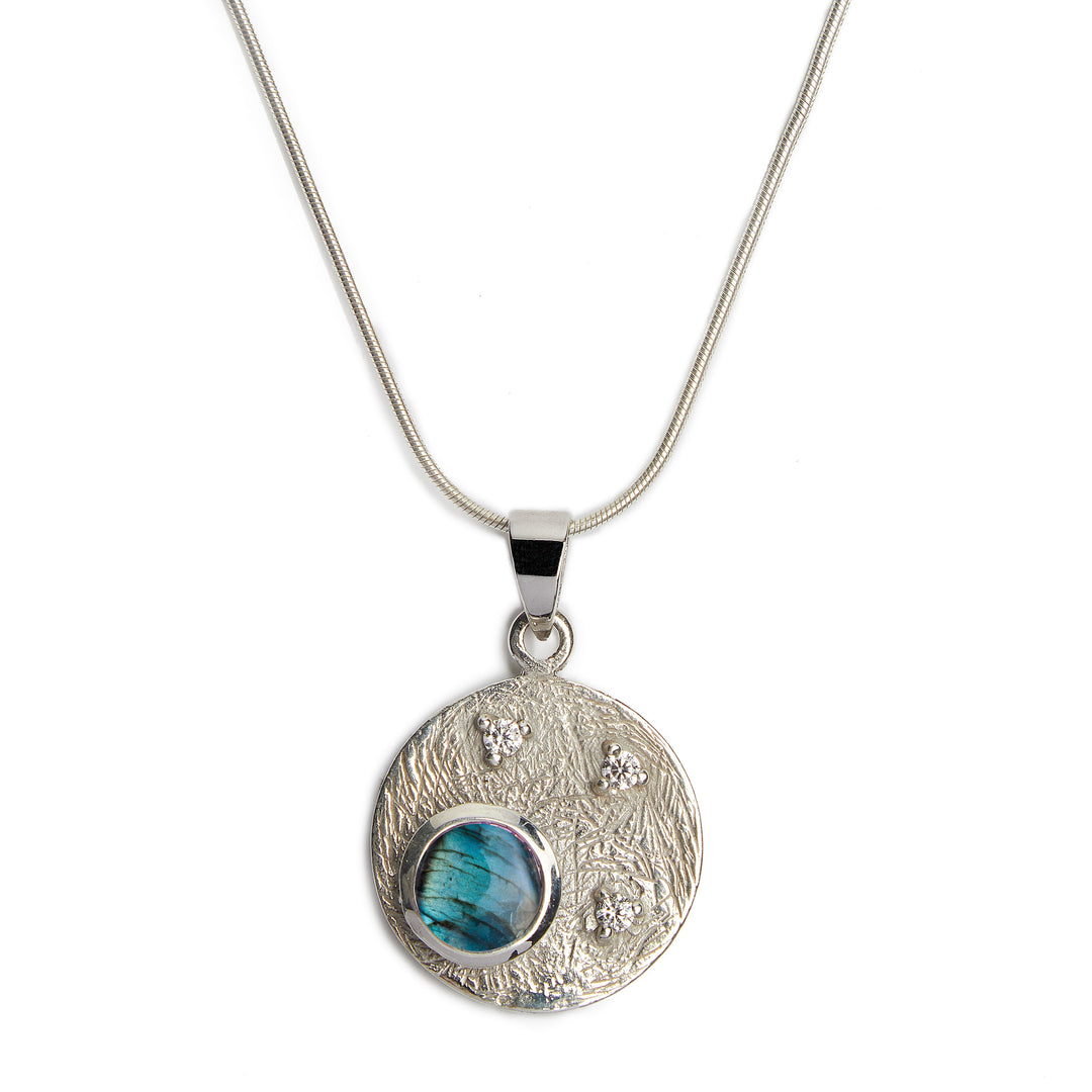 Introducing the Celeste Necklace in Silver & various gemstones by Gallardo & Blaine Designs: A sterling silver piece featuring a round pendant with a textured surface. The pendant showcases three small, cubic zirconia stones and a larger, iridescent blue-green stone set in a circular enclosure. The necklace boasts a sleek, minimalist chain.