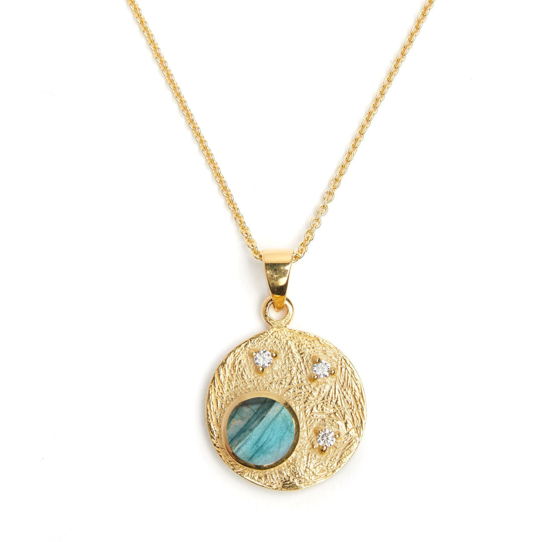 A Gallardo & Blaine Designs Celeste Necklace in Gold & various gemstones with a textured circular pendant featuring a blue-green gemstone in the center, accented by three small white cubic zirconia stones arranged in a triangular pattern. The stunning gemstone pendant hangs elegantly from a fine gold chain.