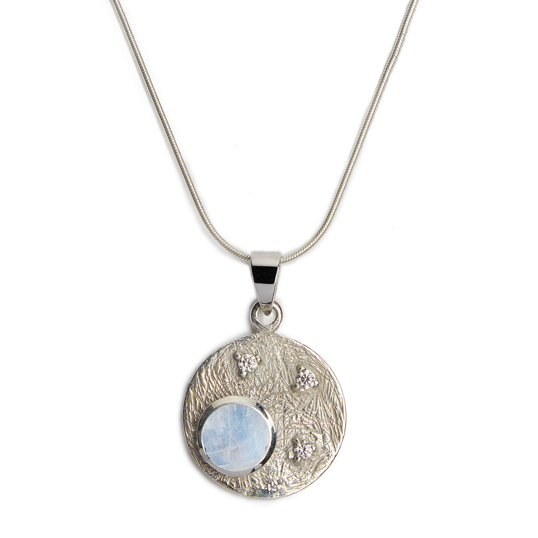 Introducing the Celeste Necklace in Silver & various gemstones by Gallardo & Blaine Designs, a sterling silver pendant necklace featuring a round, textured disc with a polished finish. The disc displays a translucent blue gemstone and three smaller sparkling cubic zirconia stones in a celestial-inspired design, all hung on a sleek snake chain.