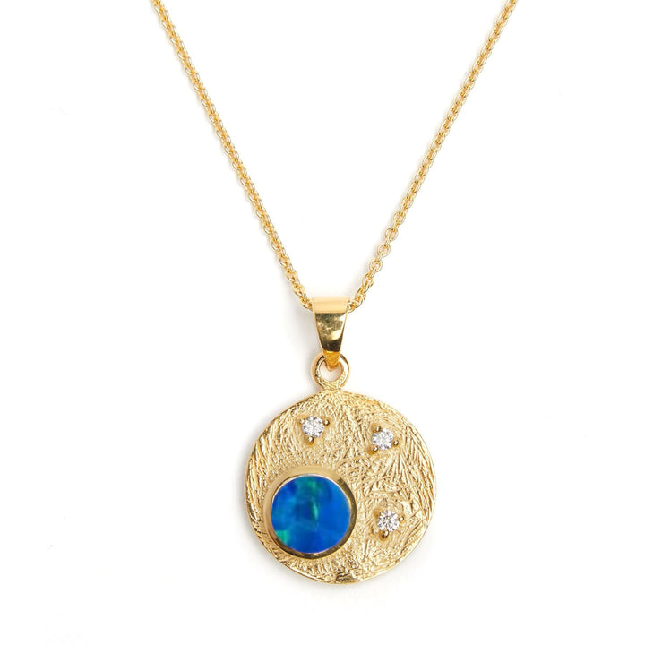 A Gallardo & Blaine Designs Celeste Necklace in Gold & various gemstones with a textured circular gemstone pendant featuring a round blue gemstone and three small clear cubic zirconia stones is shown on a white background. The pendant hangs from a delicate gold chain.